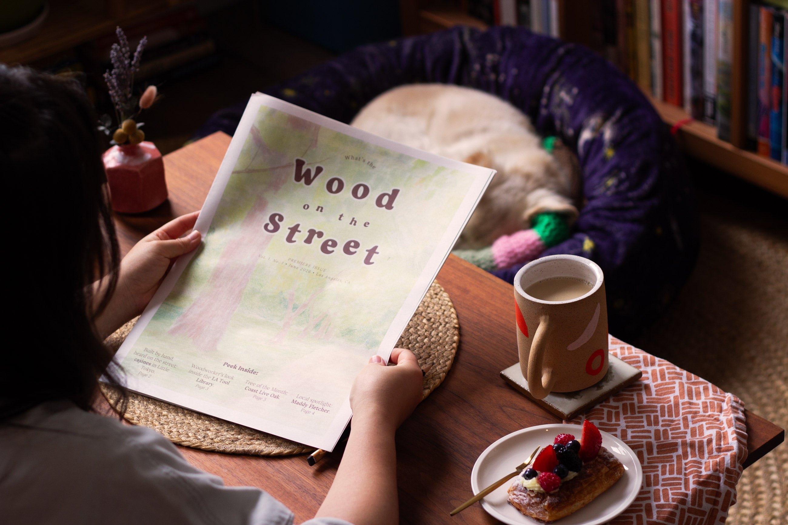 A girl holding a Wood on the Street newspaper is sitting at a wooden table, with a mug and a pastry next to her. On the floor is a sleeping dog.