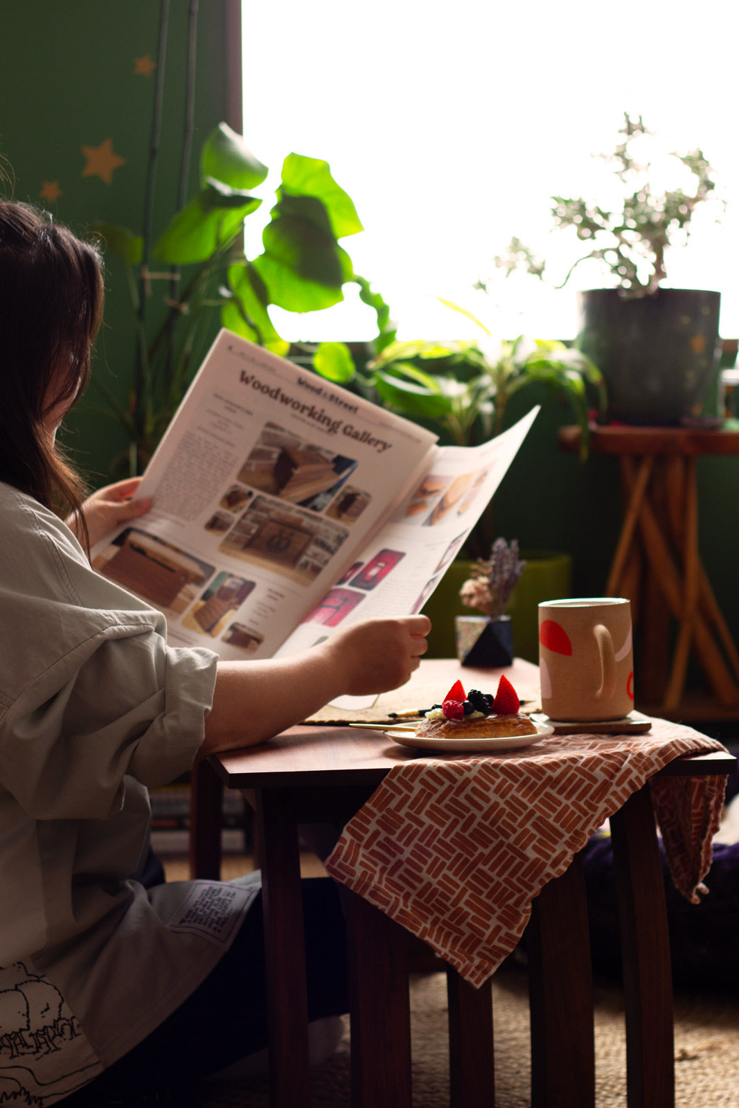 Person reading a newspaper at a wooden table with plants in the background