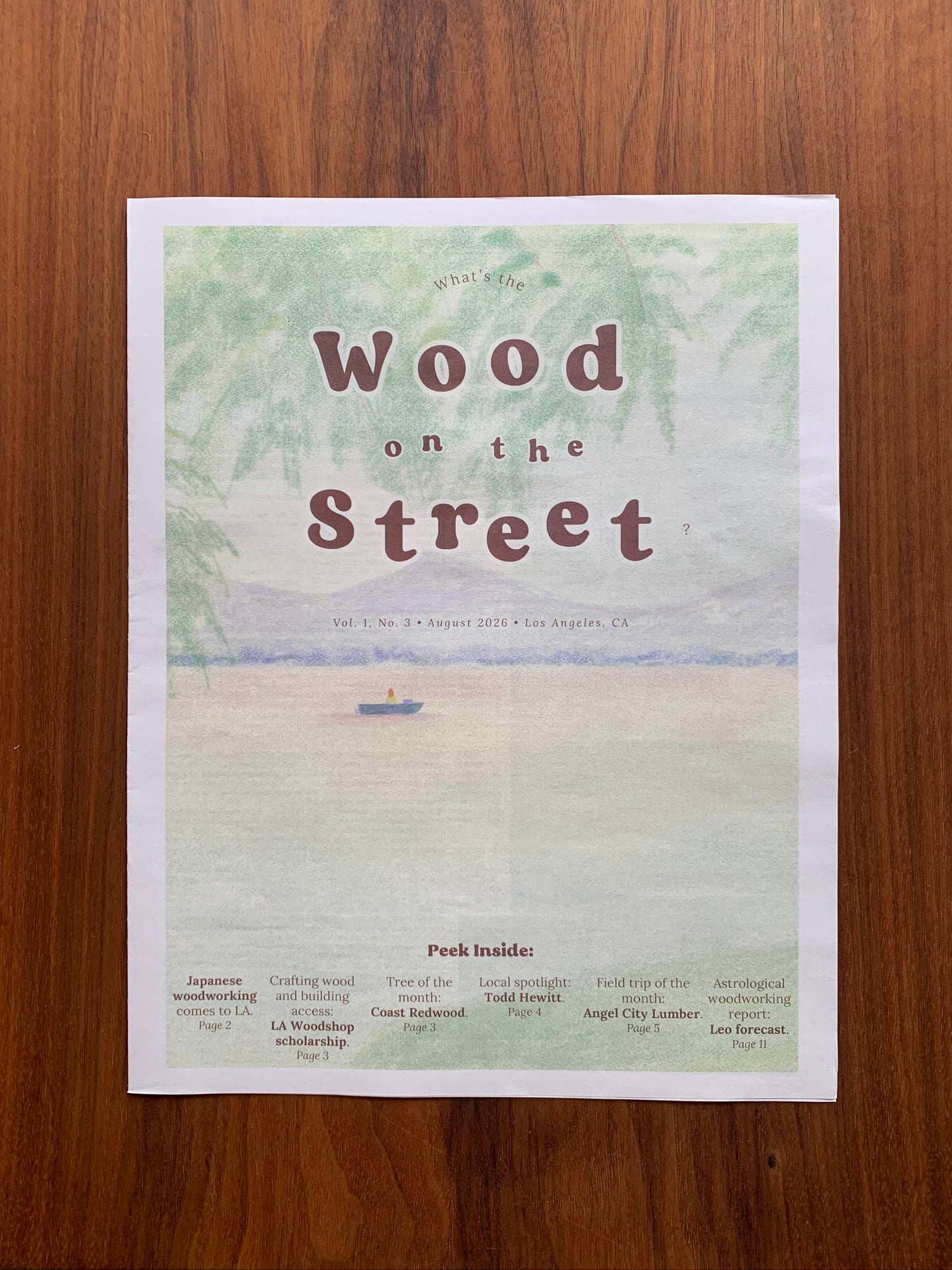 A printed newspaper titled 'Wood on the Street' with a colored pencil-style beach illustration, photographed flat on a wooden surface.