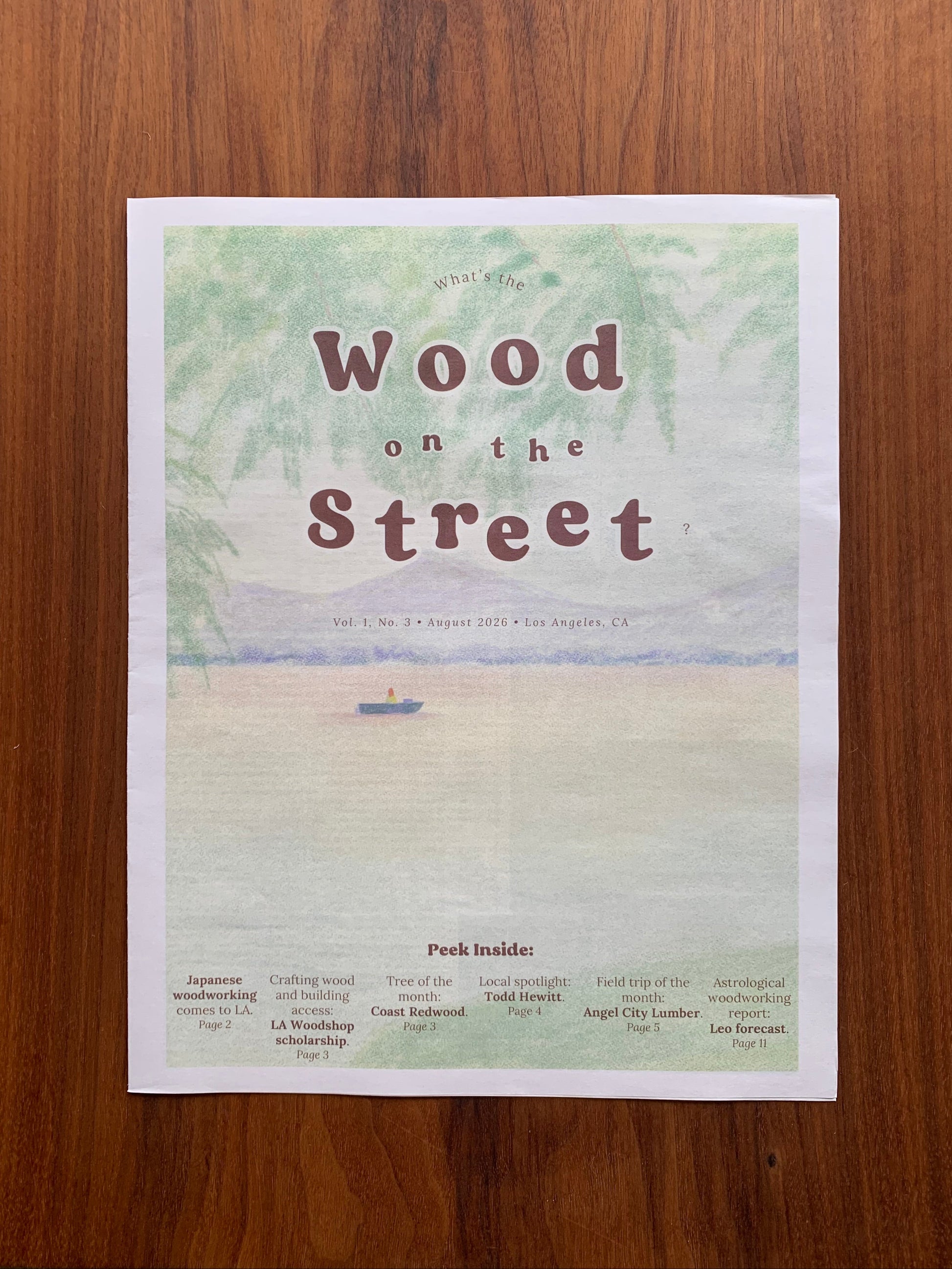 A printed newspaper titled 'Wood on the Street' with a colored pencil-style beach illustration, photographed flat on a wooden surface.